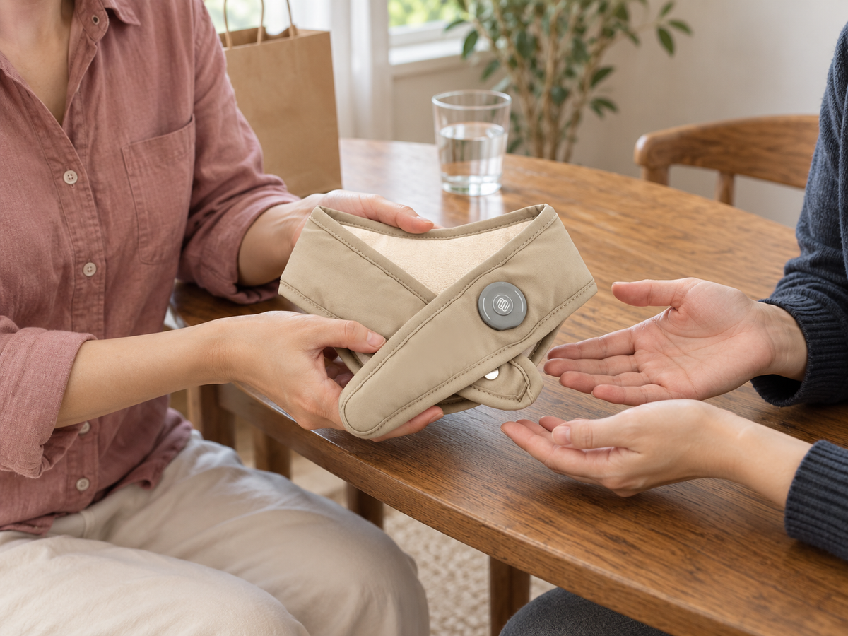 
Gifting illustration: two adults pass a champagne beige OmmiCare Neck Warmer 2.0 across a table.